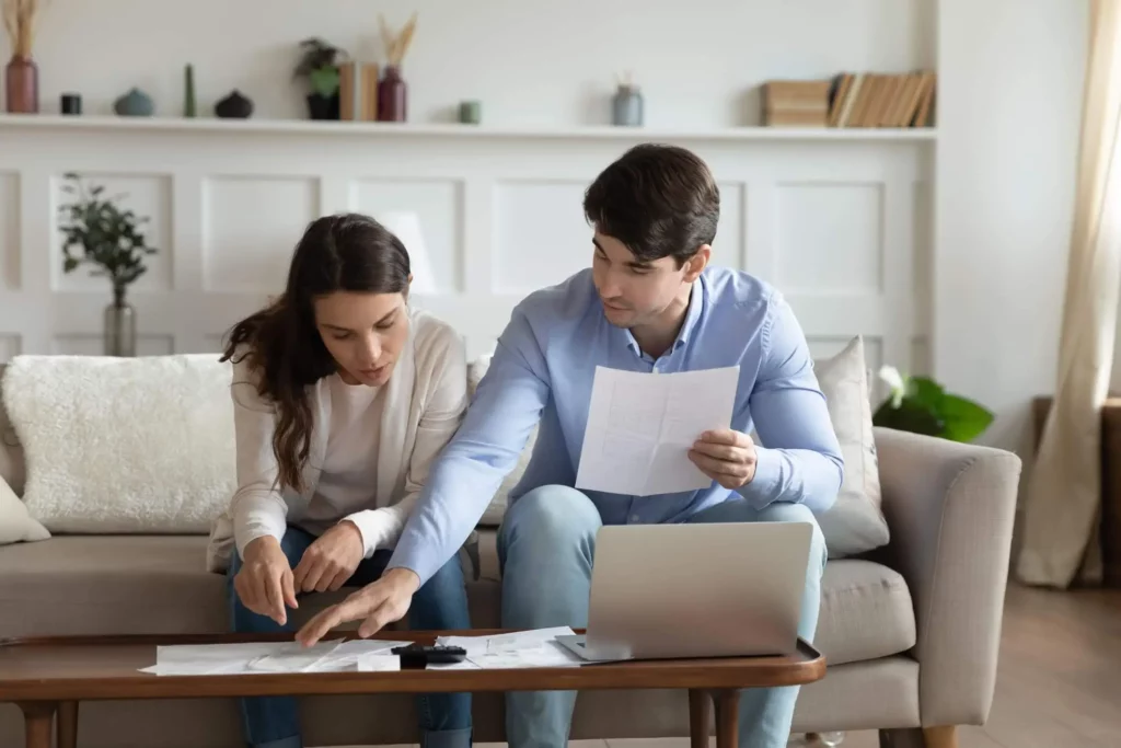 Imagem de um casal sentado em um sofá, olhando para documentos espalhados em uma mesa de centro. A mulher aponta para um documento, enquanto o homem segura outra folha e observa. Um notebook está aberto na frente deles