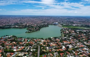 Vista aérea dos bairros da região da Pampulha, em Belo Horizonte, com a lagoa cercada por casas, árvores verdes e prédios urbanos sob um céu azul parcialmente nublado.
