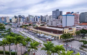 Vista aérea de uma rua movimentada da cidade, ladeada por palmeiras e tráfego, com um grande edifício histórico amarelo em frente a prédios modernos sob um céu nublado.