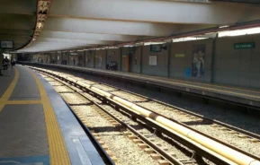 Uma plataforma de estação de trem coberta com pavimentação tátil amarela passa ao lado de dois conjuntos de trilhos de trem; a estação parece vazia, exceto por algumas pessoas ao longe. As placas na parede indicam ser o metrô São Cristóvão.