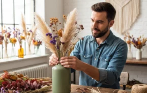 Um homem de camisa jeans arruma flores secas, incluindo grama dos pampas e flores roxas, em um vaso verde sobre uma mesa de madeira em uma sala iluminada e aconchegante, decorada com arranjos florais e arte em macramê.