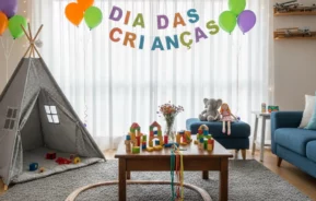 Uma sala de estar decorada com balões, uma faixa onde se lê DIA DAS CRIANÇAS, uma tenda de brincar, brinquedos, um trem de madeira, animais de pelúcia e blocos coloridos sobre uma mesa e um tapete, comemorando o Dia das Crianças.