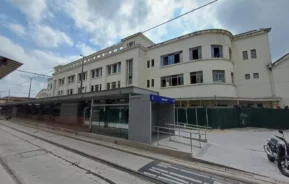 Parada de bonde moderna chamada Mercado com uma rampa e um abrigo em frente ao grande edifício branco do Mercado Municipal de Santos. Os trilhos do bonde e uma motocicleta são visíveis, sob um céu azul nublado.