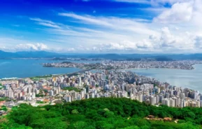 Foto que ilustra matéria sobre a Região Metropolitana de Florianópolis mostra uma parte da Capital Catarinense vista do alto, com vários edifícios altos, cercada por água e colinas verdejantes, sob um céu azul com nuvens dispersas.