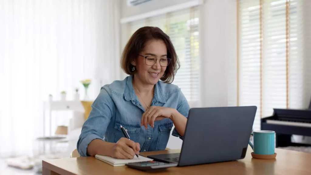 Imagem de uma mulher usando óculos e uma camisa jeans sorrindo enquanto trabalha em um laptop em uma mesa de madeira, segurando uma caneta e escrevendo em um caderno. Uma calculadora e uma caneca também estão sobre a mesa.