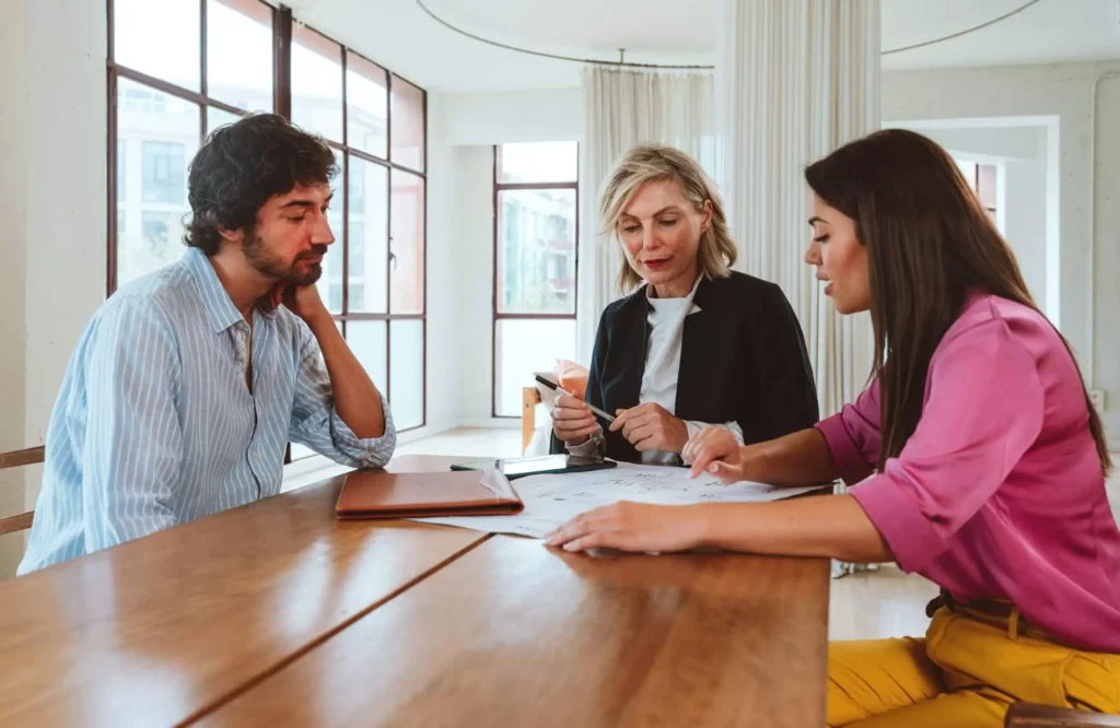 Imagem de três pessoas sentadas em uma mesa de madeira em um escritório bem iluminado, discutindo documentos. Uma mulher de blazer segura papéis, enquanto os outros dois, um homem e uma mulher, ouvem e analisam os documentos juntos.