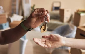 Imagem em close-up de uma pessoa entregando chaves de casa com um chaveiro em forma de casa para outra pessoa, com caixas móveis visíveis no fundo desfocado.