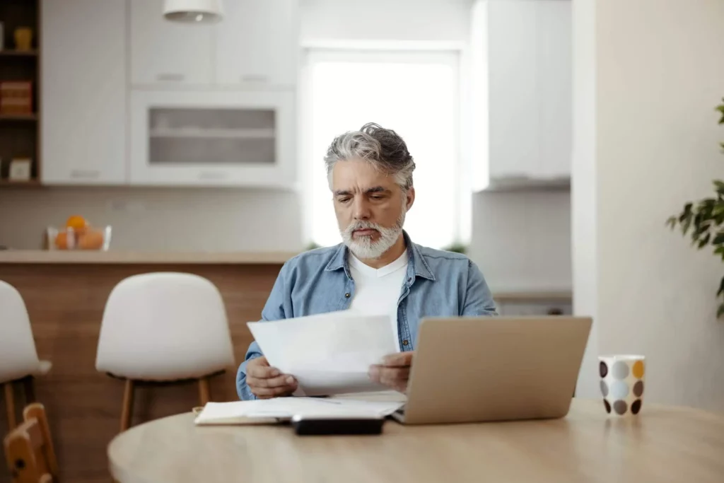 Imagem de um homem mais velho, com cabelos grisalhos e barba, sentado à mesa da cozinha, olhando para papéis em suas mãos. Um notebook, um bloco de notas e uma caneca estão sobre a mesa à sua frente. A cozinha é clara e moderna.