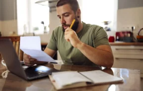 Imagem de um homem de barba sentado em uma mesa de cozinha, olhando atentamente para uma folha de papel enquanto segura um lápis. Um laptop aberto e cadernos de anotações estão sobre a mesa em frente a ele.