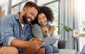 Imagem de um casal sorridente sentado junto em um sofá, olhando para um tablet. Há papéis e uma caneca na mesa à frente deles, e a luz do sol atravessa as grandes janelas ao fundo