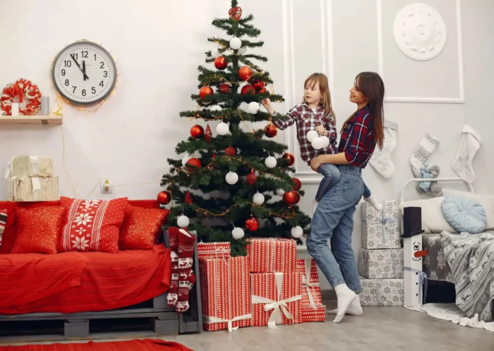 Imagem de uma mulher e uma menina decorando uma árvore de Natal com enfeites em uma sala de estar aconchegante e festiva.
