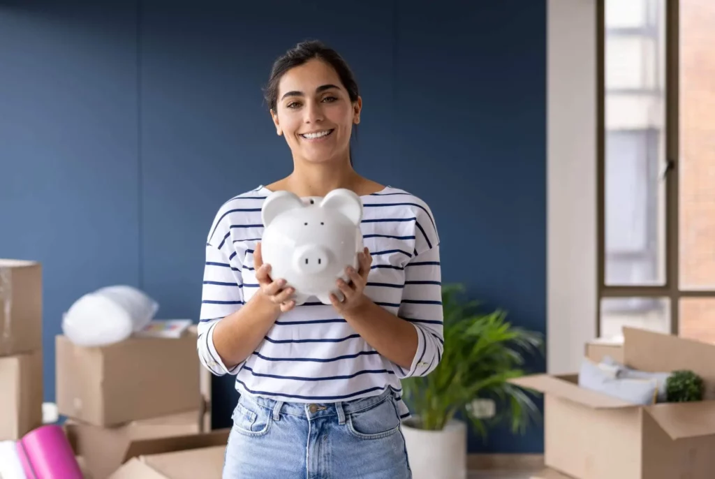 Imagem de uma mulher sorridente, com uma camisa listrada, segurando um cofrinho branco. Ela está em uma sala com caixas de mudança, um vaso de planta verde e grandes janelas ao fundo.