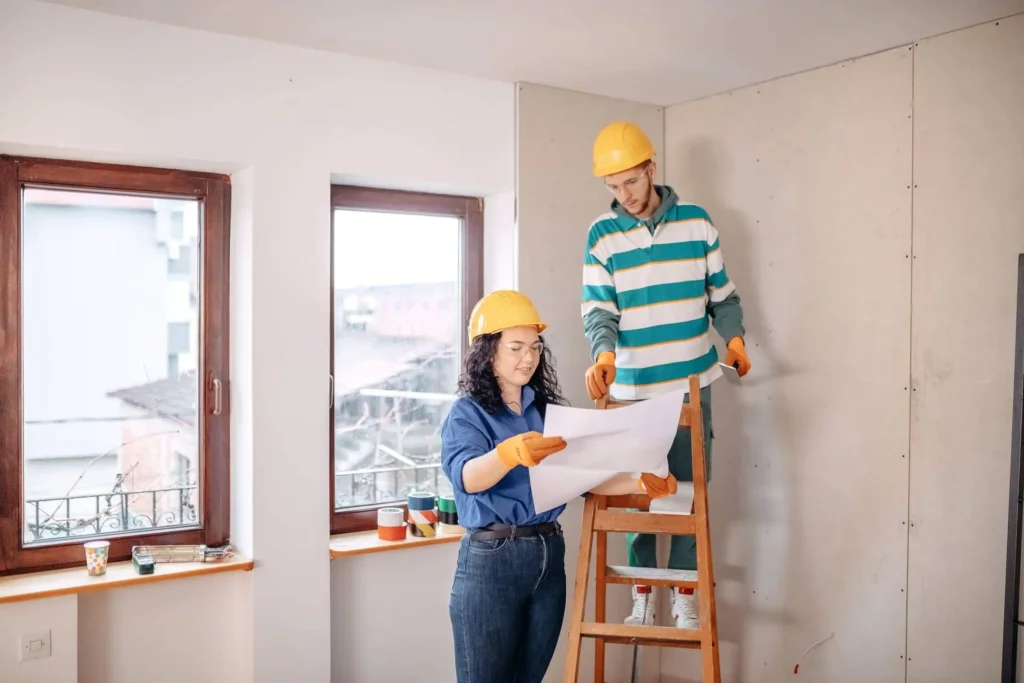 Imagem de duas pessoas usando capacetes e luvas analisando as plantas dentro de uma sala iluminada que está sendo reformada; uma delas está em uma escada, enquanto a outra segura as plantas perto de grandes janelas.