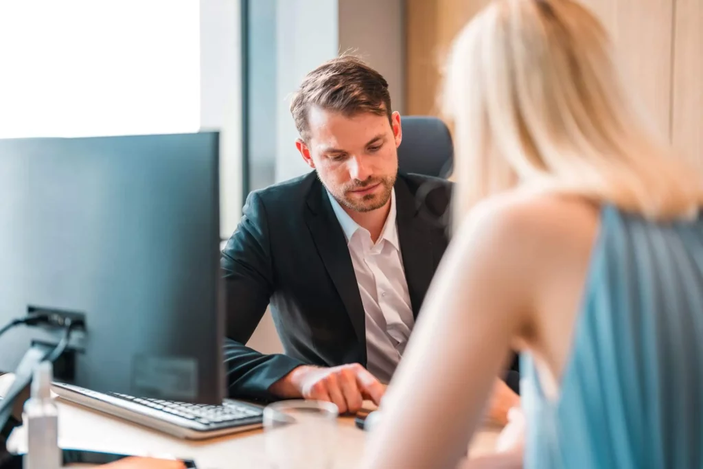 Imagem de um homem de terno sentado em uma mesa com um computador, olhando para documentos, enquanto uma mulher de cabelos loiros está sentada em frente a ele, parcialmente fora de foco.
