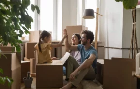 Imagem de uma família sorridente de três pessoas sentada no chão, cercada por caixas de mudança. A menina e p pai dão um high-five um no outro enquanto a mãe sorri, tudo isso em uma sala bem iluminada com grandes janelas e plantas.