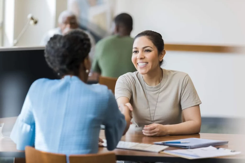 Imagem de duas mulheres sentadas em uma mesa, sorrindo e apertando as mãos, com documentos e um notebook à sua frente. Outras pessoas estão desfocadas ao fundo em um ambiente de escritório moderno e iluminado.