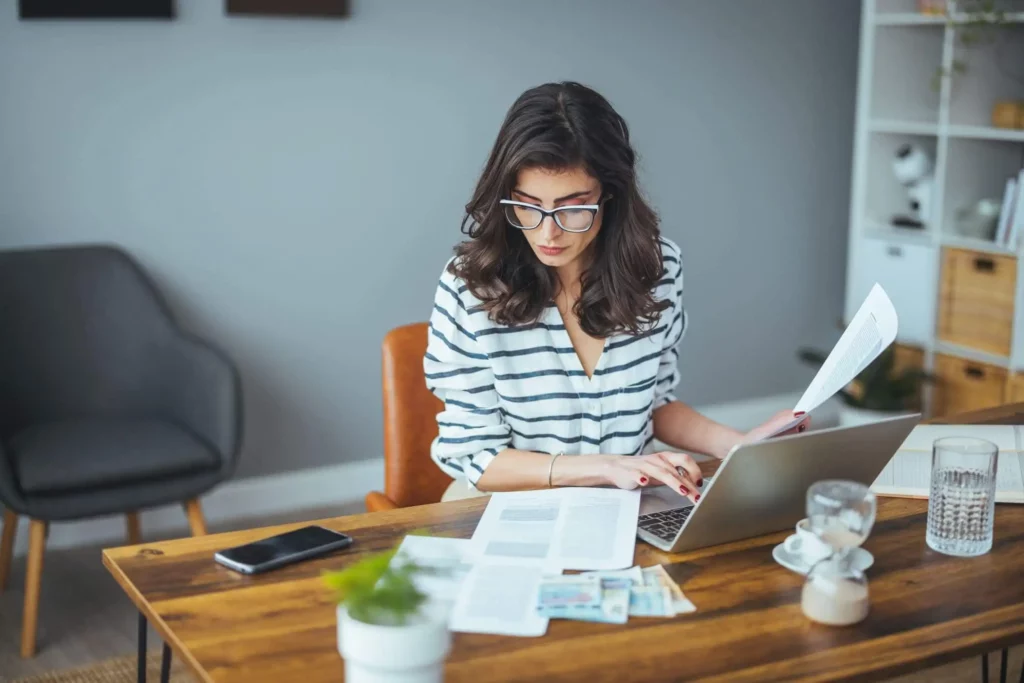Imagem de uma mulher de óculos sentada em uma mesa de madeira com um laptop, papéis, telefone e xícara de café, parecendo concentrada enquanto trabalha e revisa documentos em um escritório doméstico moderno e organizado.
