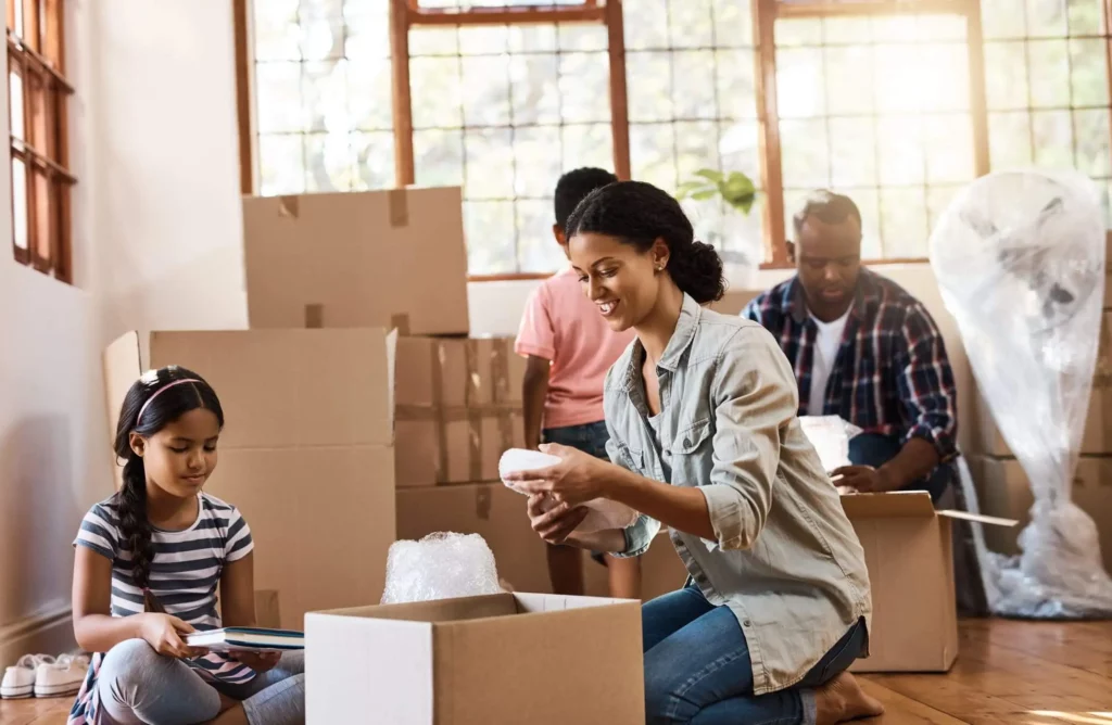 Imagem de uma família de quatro pessoas desempacotando caixas em uma sala iluminada pelo sol durante uma mudança. Uma mulher adulta desembrulha um item, uma menina segura livros e um homem e um menino organizam caixas ao fundo. Caixas de papelão estão espalhadas.