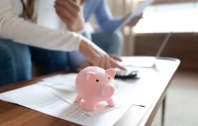 Imagem de um cofrinho rosa sobre uma mesa coberta de papéis, enquanto duas pessoas ao fundo examinam documentos e usam uma calculadora