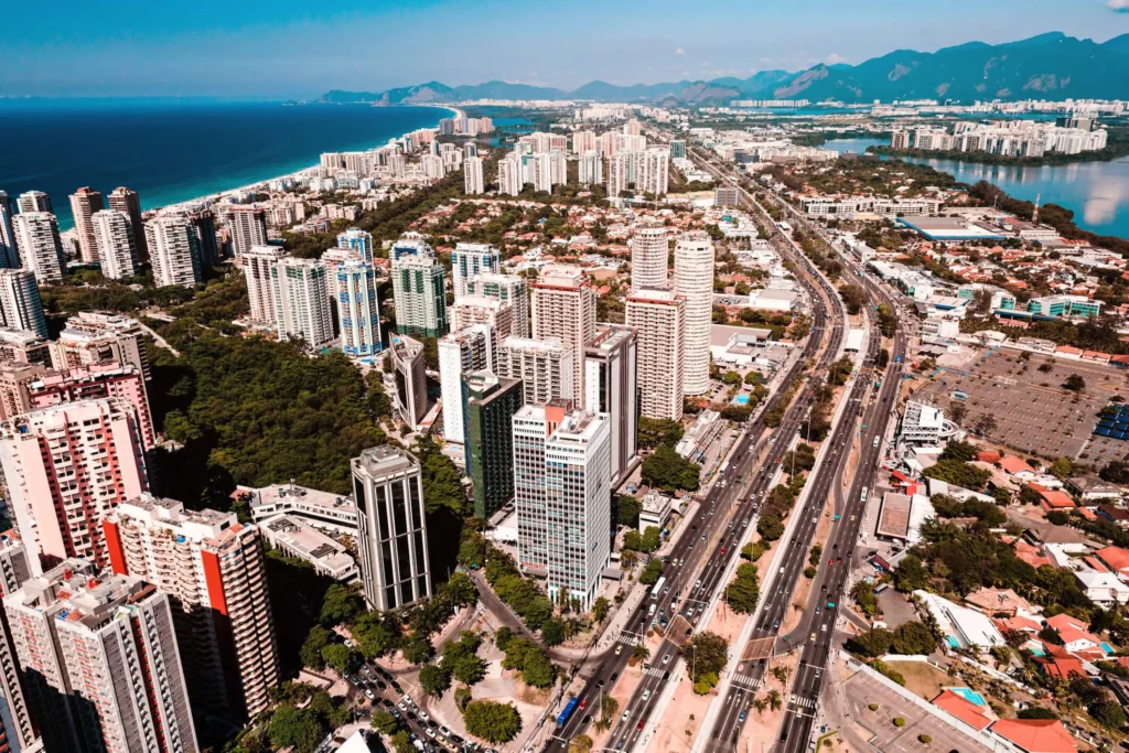 Imagem da vista aérea da Barra da Tijuca, no Rio de Janeiro, mostra edifícios altos, estradas largas, vegetação exuberante e montanhas ao fundo, ao lado de um oceano azul sob um céu claro