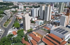 Imagem da vista aérea da cidade de Americana, SP, mostra altos prédios de apartamentos, estruturas comerciais e casas residenciais com telhados vermelhos, cortada por uma estrada principal com tráfego leve e vegetação espalhada.
