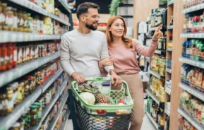 Imagem de um homem e uma mulher sorrindo enquanto fazem compras em um corredor de um supermercado. A mulher aponta para os produtos na prateleira enquanto o homem empurra um carrinho cheio de mantimentos, incluindo um abacaxi e folhas verdes.