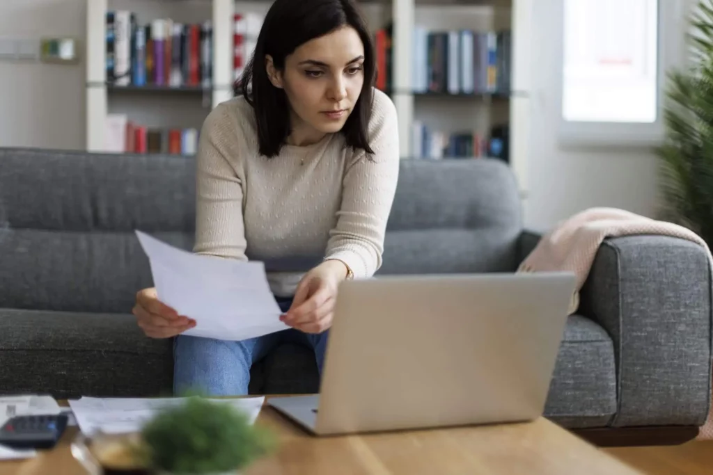 Imagem de uma mulher sentada em um sofá cinza, olhando para um laptop enquanto segura papéis. Ela parece concentrada, com estantes de livros e uma janela ao fundo. Uma mesa de centro com documentos e uma planta está em primeiro plano