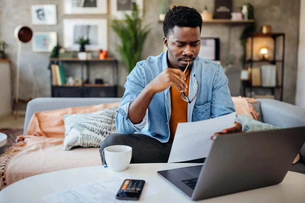 Imagem de um homem sentado em um sofá em casa, segurando óculos e olhando atentamente para um documento. Um laptop, uma caneca de café, uma calculadora e papéis estão sobre a mesa à sua frente. A sala tem uma decoração moderna e aconchegante