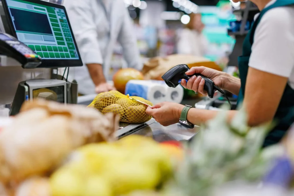 Imagem de uma pessoa no caixa de um mercado escaneando mantimentos, incluindo um saco de batatas, em um caixa de um supermercado. Vários itens alimentares estão na esteira transportadora e uma tela de registro é visível ao fundo.