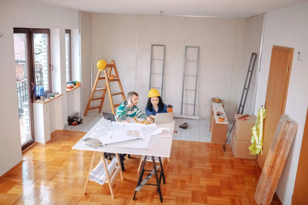Imagem de uma mulher e uma criança usando capacetes de construção sentados em uma mesa com plantas e um laptop em uma sala parcialmente reformada, cercada por ferramentas, escadas e materiais de construção.