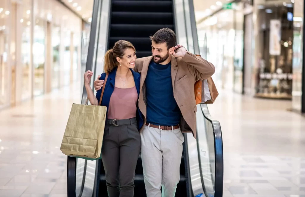 Imagem de um casal sorridente segurando sacolas de compras em uma escada rolante em um shopping center bem iluminado, olhando um para o outro com carinho