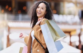 Imagem de uma mulher sorridente com cabelos cacheados segurando várias sacolas de compras coloridas no ombro enquanto caminha ao ar livre em um shopping, vestida com um casaco marrom.