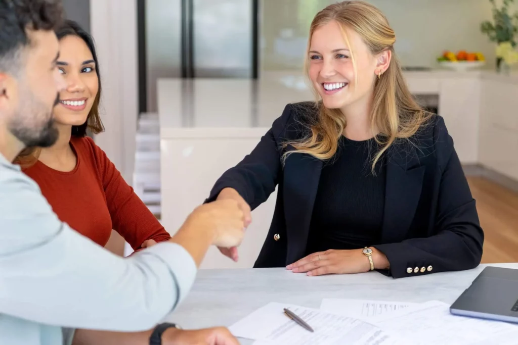 Imagem de uma mulher sorridente, vestindo um blazer preto, cumprimentando um homem em uma mesa, enquanto outra mulher observa e sorri. Papéis e uma caneta estão sobre a mesa em um ambiente de escritório moderno e iluminado