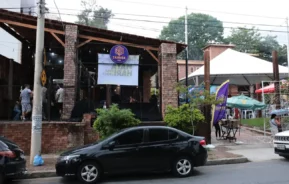 Um carro preto está estacionado em uma rua em frente a uma cafeteria de tijolos com uma grande placa onde se lê "Chakara". As pessoas podem ser vistas dentro e fora da cafeteria, e guarda-chuvas coloridos cobrem as áreas de assentos ao ar livre.