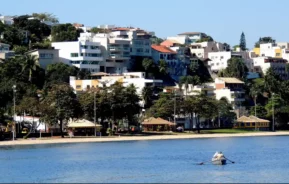 Uma pessoa rema um pequeno barco em águas azuis calmas perto da costa, com um pano de fundo de casas modernas, brancas e coloridas, em uma pequena colina exuberante e coberta de árvores, sob um céu claro, onde fica a Bairros da Ilha do Governador - Praia da Bica, no Jardim Guanabara, um dos bairros da Ilha do Governador, no Rio de Janeiro.