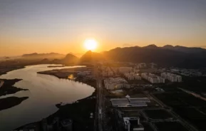 Vista aérea de uma cidade ao pôr do sol, com o sol se pondo atrás das montanhas. A lagoa de Jacarepaguá aparece lado de edifícios e ruas urbanas, lançando longas sombras e criando um brilho dourado na paisagem.