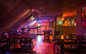 Um bar pouco iluminado com mesas e cadeiras de madeira escura, iluminação de neon colorida, pôsteres nas paredes e um teto em estilo industrial. A atmosfera é aconchegante e convidativa, com uma vibração moderna e urbana.