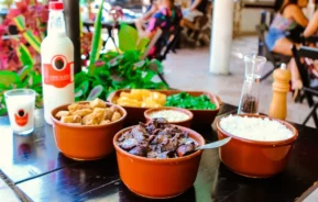 Uma mesa posta com tigelas de comida brasileira, incluindo arroz, ensopado de carne, couve, farofa, laranjas fatiadas e uma garrafa de cachaça, com pessoas sentadas ao fundo em um restaurante ao ar livre.