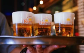 Uma pessoa segura uma bandeja com três copos de cerveja, cada um com o logotipo de Redentor, um dos Bares da Savassi, com uma espuma espessa, em um ambiente interno com iluminação quente.