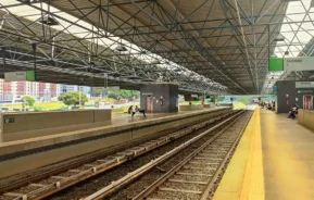 Estação Águas Claras do Metrô-DF: moderna e ao ar livre com plataformas cobertas, trilhos de trem passando pelo centro e algumas pessoas esperando. O teto tem uma estrutura de metal e os edifícios da cidade são visíveis ao fundo.