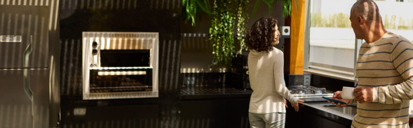 Um homem segurando uma caneca e um tablet está perto de uma mulher em uma cozinha moderna com bancadas pretas, um forno embutido e uma grande planta. A luz do sol entra pelas janelas.