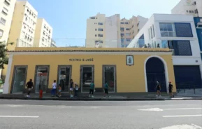Fachada do Mercadinho São José, localizado em uma rua da cidade com pessoas caminhando, prédios de apartamentos altos e estruturas modernas visíveis ao fundo.