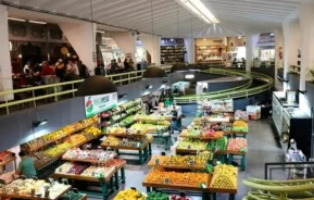 Parte interna do Mercado Municipal de Pinheiros, um lugar espaçoso com barracas bem organizadas exibindo frutas e legumes coloridos. As pessoas fazem compras e caminham enquanto outras se sentam em mesas em um nível superior. O espaço apresenta grandes luzes suspensas e uma varanda curva.