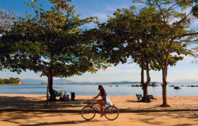 Uma mulher anda de bicicleta em uma trilha de areia na praia, sombreada por árvores, com pessoas relaxando sob as árvores e um mar calmo com rochas ao fundo, sob um céu azul claro.