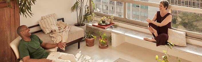 Um homem e uma mulher estão sentados um em frente ao outro em uma sala de estar moderna e iluminada, com grandes janelas e vários vasos de plantas, sorrindo e conversando.