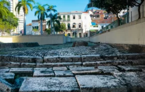 Degraus de pedra levam a uma praça com palmeiras, edifícios em estilo colonial e um céu azul claro ao fundo. Prédios urbanos na encosta são visíveis à direita. O cenário é o Cais do Valongo, que faz parte da região conhecida como Pequena África, no Rio de Janeiro.