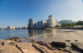 Uma praia de areia com grandes rochas em primeiro plano, ondas calmas e uma fileira de edifícios altos e modernos ao longo da costa sob um céu azul claro. Árvores e montanhas distantes são visíveis ao fundo.
