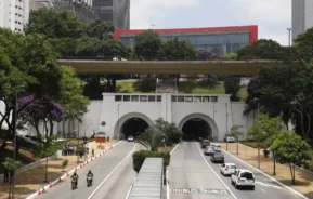 Imagem da entrada do Túnel 9 de Julho, em São Paulo, mostra a entrada para veículos, um viaduto para pedestres acima, árvores em ambos os lados e o MASP atrás. Pessoas e ciclistas podem ser vistos perto da estrada.