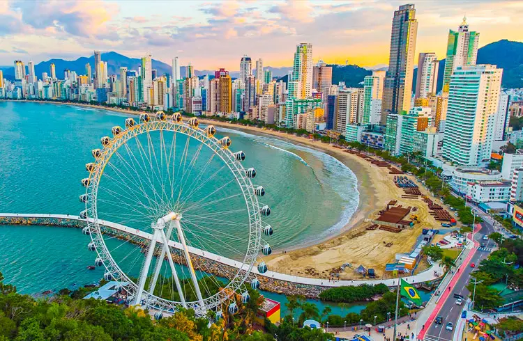 Imagem de uma grande roda-gigante perto de uma praia com águas calmas e azuis em Balneário Camboriú, Santo Catarina, cercada por arranha-céus altos e modernos. A paisagem urbana é vibrante e o pôr do sol lança um brilho quente sobre a área urbana costeira.
