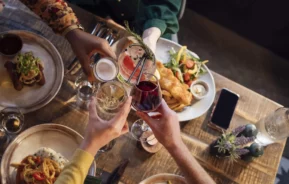 Imagem de quatro pessoas brindando com bebidas diferentes em uma mesa de madeira com pratos de comida, incluindo salada, massa e peixe frito, criando uma atmosfera calorosa e comemorativa.